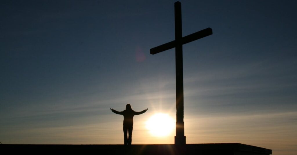 Silhouette of a person with open arms beside a cross at sunset, symbolizing faith and spirituality.